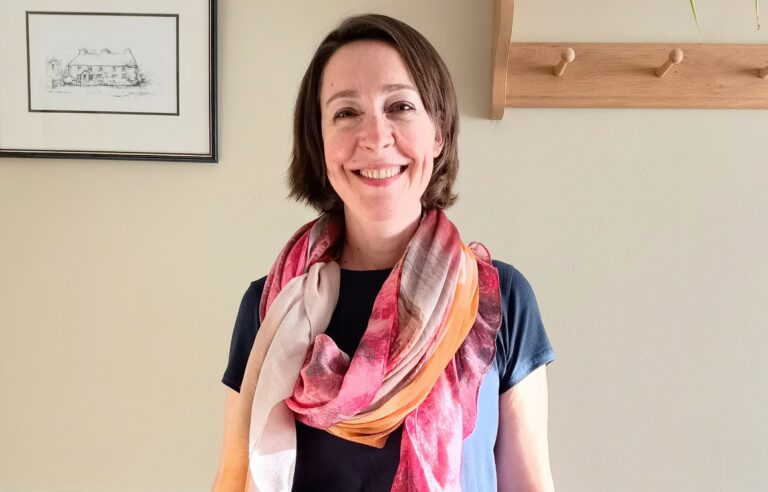 photo of Ingrid Pope, smiling, standing in front of a wall, wearing jeans, a blue t-shirt and a pink scarf